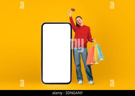 Young Woman tiene borse e carte vicino al grande smartphone Mockup in un ambiente giallo brillante Foto Stock