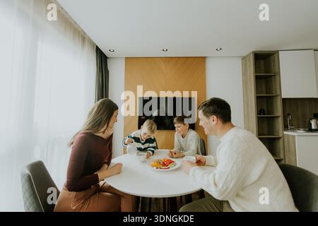 Quattro membri della famiglia siedono a un tavolo rotondo in una cucina moderna. I bambini sono impegnati a fare colazione. La luce del sole riempie la stanza, creando una calda a Foto Stock