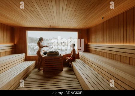 Due persone godono di una tranquilla esperienza di sauna, circondate da caldi interni in legno e da un paesaggio invernale mozzafiato all'esterno. Foto Stock