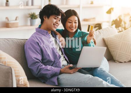Famiglia asiatica positiva che fa shopping in linea con carta di credito e laptop, seduta sul divano e coccole a casa Foto Stock