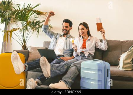 Coppia felice prenotazione hotel online con computer portatile e carta di credito, seduta con valigie, passaporti e biglietti Foto Stock