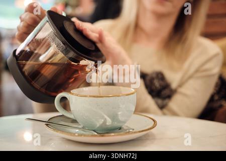 Persona che versa una bevanda calda da una pentola di vetro in una tazza di ceramica in una caffetteria, illustrando uno stile di vita accogliente e un concetto di ospitalità. Foto Stock