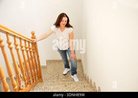 donna incinta che sale le scale a casa e gli tocca il ginocchio per il dolore Foto Stock