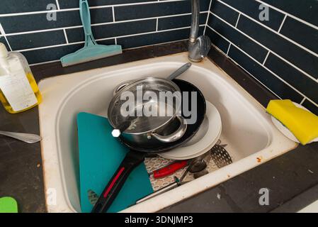 Lavandino della cucina sporco pieno di piatti non lavati, utensili e utensili per la pulizia, che illustrano il disordine domestico e le faccende domestiche di tutti i giorni. Foto Stock