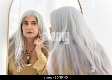 Una donna dai capelli grigi di mezza età che guarda allo specchio, tocca gli zigomi Foto Stock