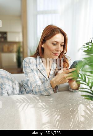 Una donna si siede su un divano in una casa mentre scrive messaggi al telefono. Sembra felice e rilassata nei pigiami durante il giorno. Foto Stock
