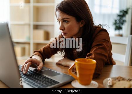 Una donna siede a un tavolo in salotto. Si concentra sul suo laptop mentre sorseggia il tè. Si sente stanca e annoiata Foto Stock