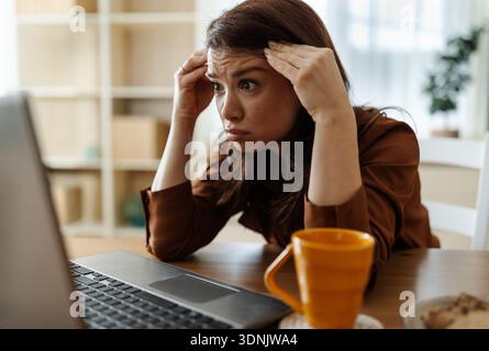 Una donna siede a un tavolo guardando il suo notebook con un'espressione preoccupata. Tiene la fronte e sembra stressata Foto Stock