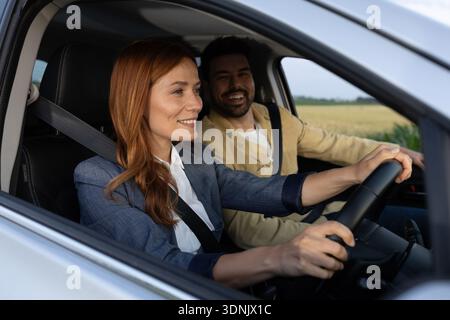 Prova a guidare una macchina nuova. Donna che guida e sorride con sicurezza mentre l'uomo sul sedile del passeggero la guarda con un sorriso sul viso e si gode la dri Foto Stock