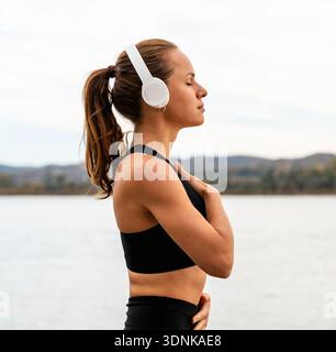 Donna attenta con le cuffie che tiene le mani sul petto facendo esercizio respiratorio nella natura vicino al fiume. Foto Stock