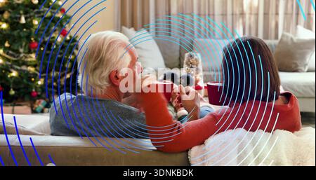 Sorseggiando una coppia senior che indossa maglioni, seduto sul divano di casa con rivestimento blu, tenendo in mano tazze rosse Foto Stock