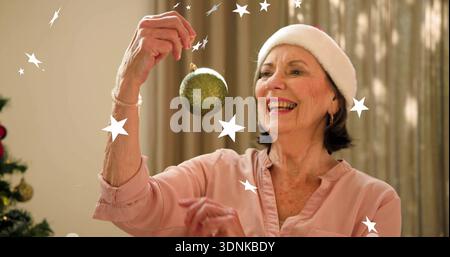 Tenendo in mano un ornamento verde, donna che ispeziona a casa, albero di vacanza, camicetta rosa, cappello di Babbo Natale Foto Stock