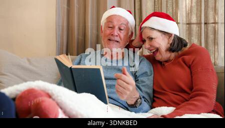 Lettura di una coppia anziana seduta sul divano del soggiorno, indossando cappelli di Babbo Natale e tenendo un libro con copertina rigida Foto Stock