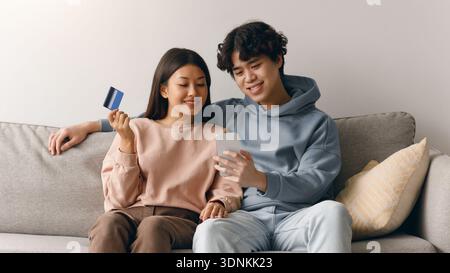Coppia seduta su un divano che guarda il telefono mentre tiene in mano una carta di credito durante lo shopping Foto Stock