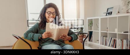 Donna che legge un libro seduto su una sedia in una stanza luminosa con scaffali e piante Foto Stock