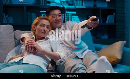 Tranquilla coppia multirazziale che guarda il cinema e la televisione coccolarsi riposando in un comodo soggiorno sul divano la sera a casa di un uomo asiatico eterogeneo Foto Stock