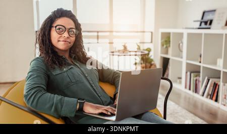 La giovane donna lavora sul computer portatile mentre si siede comodamente in una camera moderna durante le ore diurne Foto Stock