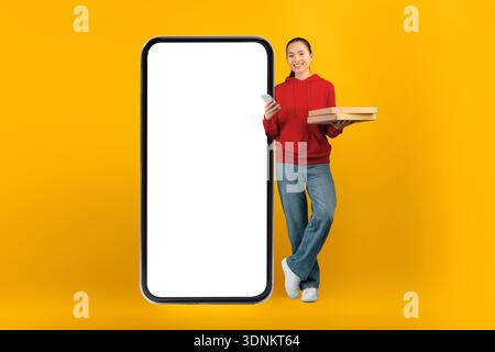 Giovane donna in maglione rosso accanto al telefono grande con scatole per la consegna di alimenti in mano Foto Stock