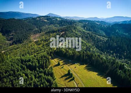 Vista aerea della lussureggiante valle verde circondata da colline boscose e montagne sotto il cielo azzurro. Campeggio con tende e veicoli annidati nella radura, con sentieri tortuosi che attraversano il paesaggio. Foto Stock