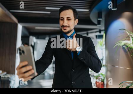 Un uomo d'affari sicuro di sé con una tuta scatta un selfie in un ufficio moderno, mostrando positività Foto Stock