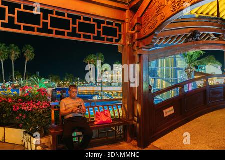 Uomo asiatico anziano che guarda uno smartphone seduto su una panchina di legno di notte a Sanya Foto Stock