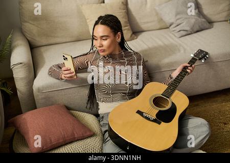 Giovane donna con trecce seduto comodamente sul pavimento, controllando il telefono mentre tiene in mano una chitarra. Foto Stock