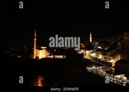 Vista notturna della storica città vecchia di Mostar con case in pietra illuminate, minareti moschea, caffetterie sul fiume e luci calde che si riflettono lungo il Ner Foto Stock