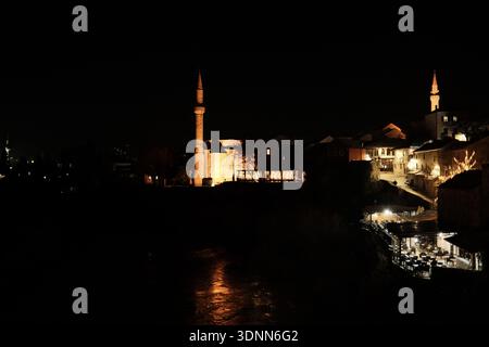 Vista notturna della storica città vecchia di Mostar con case in pietra illuminate, minareti moschea, caffetterie sul fiume e luci calde che si riflettono lungo il Ner Foto Stock