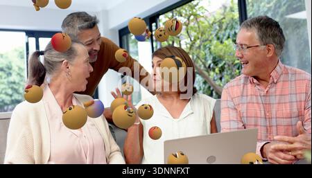 Uomo pendente sorridente mentre gli amici ridono, parlano intorno al laptop in salotto con sfere emoji Foto Stock