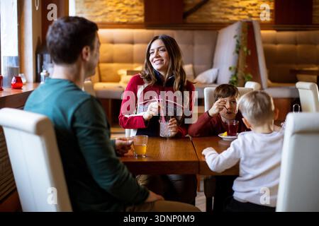 Una famiglia siede ad un tavolo di legno in un ristorante. Condividono bevande e cibo mentre sono impegnati in una conversazione. Due bambini sono seduti con loro, divertendosi Foto Stock