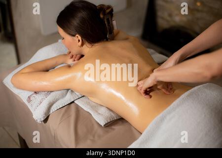 Una donna giace su un tavolo da massaggio in una spa mentre un terapista le applica l'olio sulla schiena. Il terapista usa le mani per un massaggio. Gli asciugamani sono morbidi Foto Stock