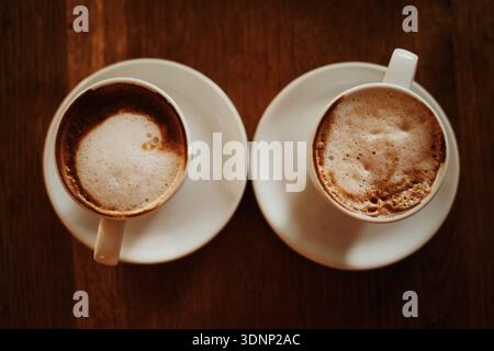 Due tazze per cappuccino sono visibili dall'alto su un tavolo in legno con una ricca consistenza in schiuma. Concetto di equilibrio, simmetria, momenti condivisi e caffè moderno Foto Stock
