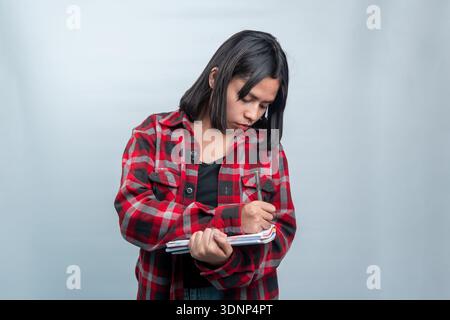 Una giovane donna del sud-est asiatico con una camicia in flanella a quadri rossa che si concentra sulla scrittura o sulla presa di appunti in un quaderno su un semplice sfondo bianco. Foto Stock