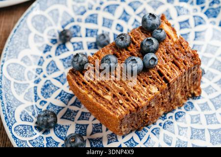 Una deliziosa fetta di torta al miele adornata con mirtilli freschi, che mostra i suoi ricchi strati. Questo dessert, popolare nella cucina Ucraina ed europea, Foto Stock