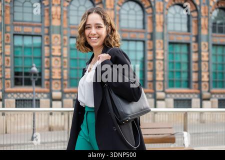 Una donna sicura di sé in un abbigliamento da lavoro cammina all'aperto, portando con sé una borsa nera mentre sorride alla fotocamera. Trasuda professionalità contro un backdr Foto Stock