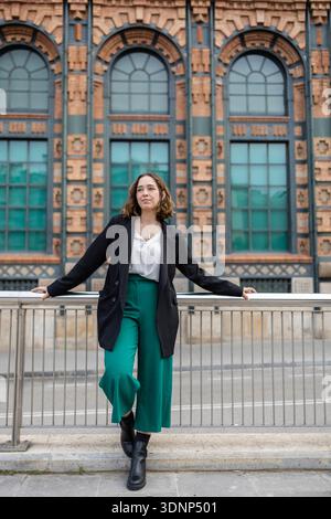 Giovane donna appoggiata su una ringhiera di metallo di fronte a un grande edificio in mattoni ornato con finestre ad arco, che indossa una giacca nera e pantaloni verdi, exud Foto Stock