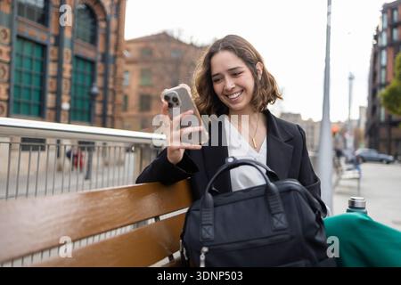 Donna seduta su una panchina in un ambiente urbano, sorridente al suo cellulare. Appare rilassata e impegnata, con una borsa e una bottiglia di bevande accanto a lei, ag Foto Stock