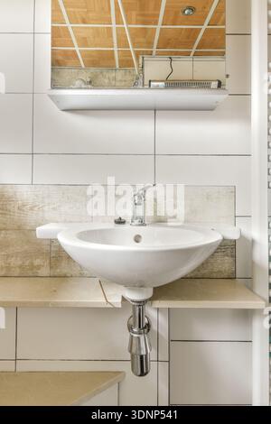 Il bagno moderno è dotato di un elegante lavandino bianco a parete con rubinetto cromato. Le tonalità neutre delle pareti piastrellate e degli scaffali in legno creano un ambiente pulito Foto Stock