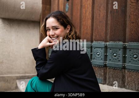 Una donna con un blazer nero e pantaloni verdi si siede comodamente su gradini di pietra, sorridendo calorosamente. La sua posa rilassata e la sua espressione allegra trasmettono un senso Foto Stock