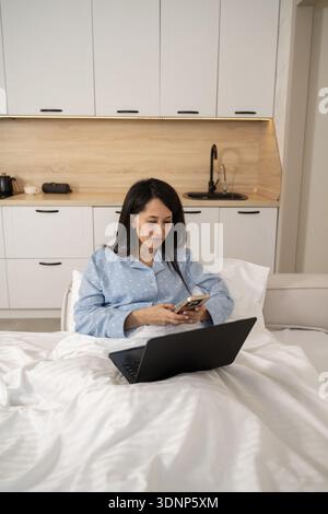 Una donna in un comodo pigiama a casa, lavora comodamente dal letto con il laptop mentre controlla lo smartphone, mettendo in risalto un mix di leisur Foto Stock