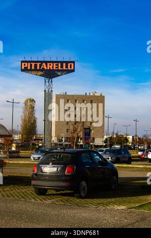 Un alto cartello commerciale Pittarello si erge sopra un parcheggio affollato con un moderno edificio in pietra sotto un cielo limpido Foto Stock