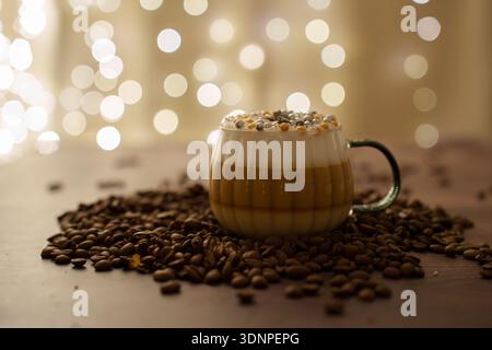 un accogliente caffè con una bevanda frizzante in una tazza di vetro posizionata su un letto di chicchi di caffè, circondata da calde luci bokeh, che creano un'atmosfera festosa e accogliente Foto Stock