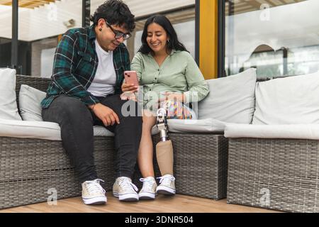 Una donna incinta con una gamba protesica e il suo partner sorridono mentre utilizza uno smartphone Foto Stock