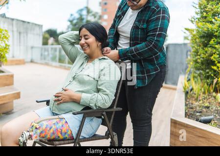 Donna incinta con una gamba protesica sorridente, che riceve assistenza dal suo partner su un balcone Foto Stock