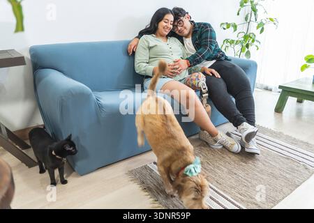 Donna incinta felice e partner con protesi gambe abbracciate sul divano a casa con animali domestici Foto Stock