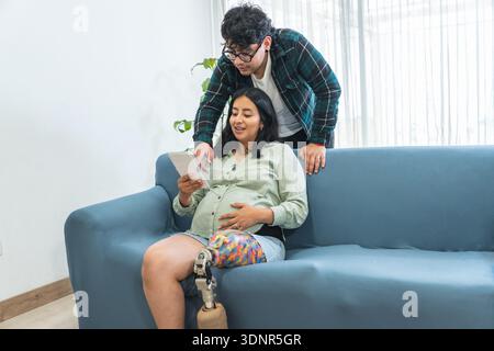 Donna amputata incinta con una gamba protesica e il suo partner che guarda un tablet digitale a casa Foto Stock