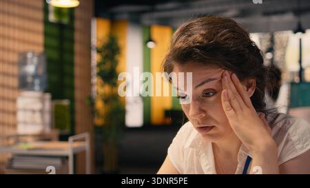 Donna d'affari che soffre mal di testa e affaticamento degli occhi, lavora in ufficio, sfregando le tempie in visibile affaticamento e stress. Emicrania e visione offuscata a causa di tensione sul lavoro, disagio. Foto Stock