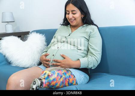 Donna incinta con una gamba protesica che sorride e tocca la pancia mentre è seduta su un divano Foto Stock