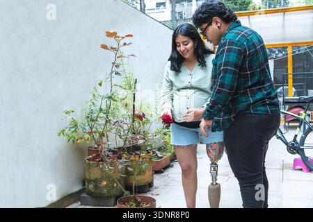 Donna incinta con una gamba protesica e il suo partner sorridente, guardando una rosa in vaso Foto Stock
