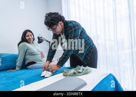 Una donna incinta felice che guarda il partner organizza gli articoli per il bambino sul letto Foto Stock